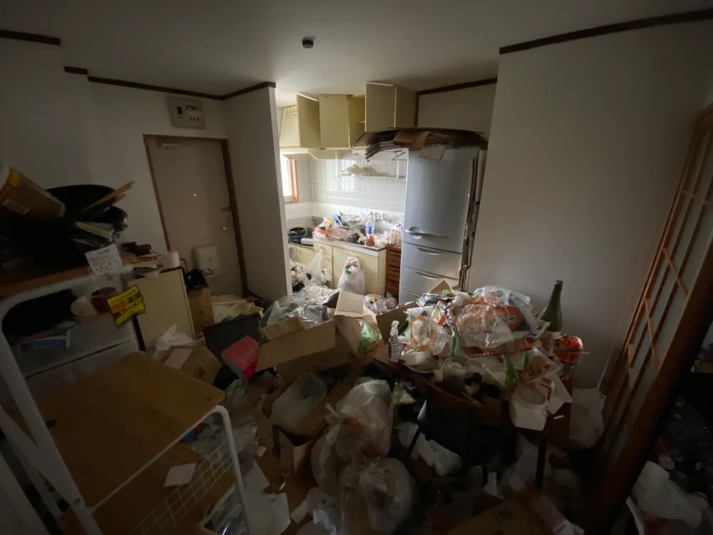 福岡県のゴミ屋敷片付け・汚部屋片付け | 九州片付け隊 福岡市南区の2DKアパートで、玄関とキッチンの床に段ボールや袋入りの生活ごみが重なり、大型冷蔵庫が残る作業前の状態